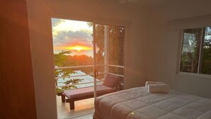 Ein Schlafzimmer mit einem großen Bett, Blick auf den Sonnenuntergang durch eine Glastür und einem Balkon mit einem Stuhl.