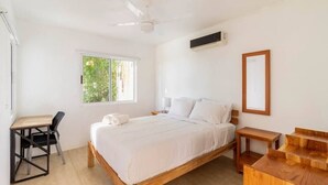 Ein Schlafzimmer mit Bett, Schreibtisch, Stuhl, einer Holzkommode, einem Deckenventilator und einem Fenster mit Blick auf Grünflächen.
