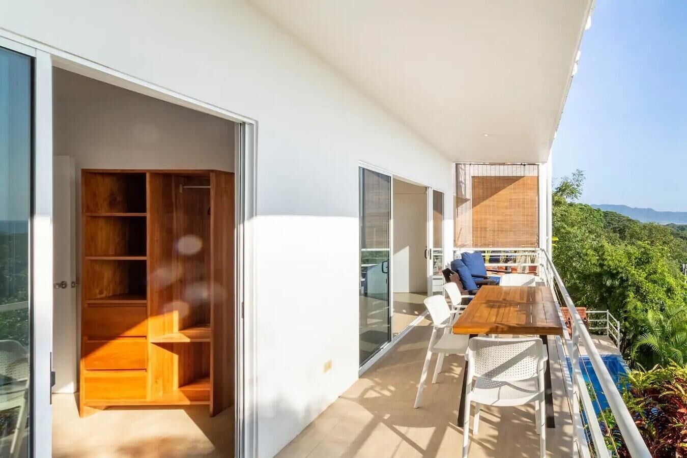 A modern balcony with a wooden table and chairs, a glass door leading to a room with wooden furniture, and a view of lush greenery.