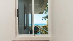 Une pièce comprenant un bureau et une chaise, une grande fenêtre donnant sur la mer et un balcon avec une balustrade.