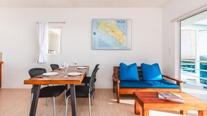 Une pièce moderne avec une table à manger en bois, dressée pour deux personnes, un canapé avec des coussins bleus et une grande fenêtre offrant une vue dégagée.