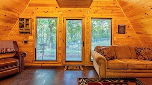 Interior acogedor de una cabaña con paredes de madera, dos sofás, una chimenea y un ventanal grande con vista a un bosque.