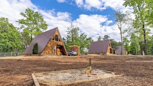 Un camping con dos cabañas de madera y una estructura central, rodeado de árboles y cielo despejado.