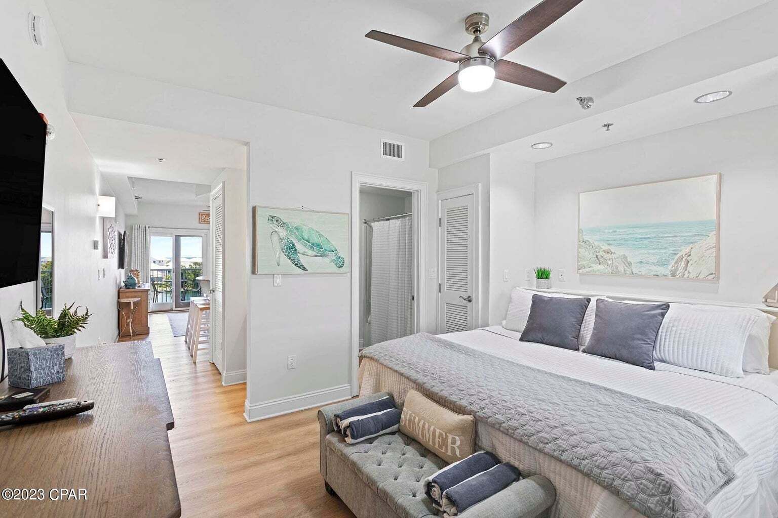 A bedroom with a large bed, a ceiling fan, a painting of a turtle, and a view of the ocean through a window.