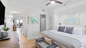 A bedroom with a large bed, a ceiling fan, a painting of a turtle, and a view of the ocean through a window.