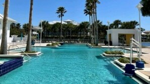 A clear blue pool surrounded by palm trees and a white building with a covered seating area.