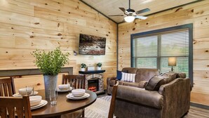 A cozy living room with a fireplace, wooden walls, a sofa, and a dining table set for four. - Cozy 2BR | HOT TUB | Pets Welcome! (Sevierville)