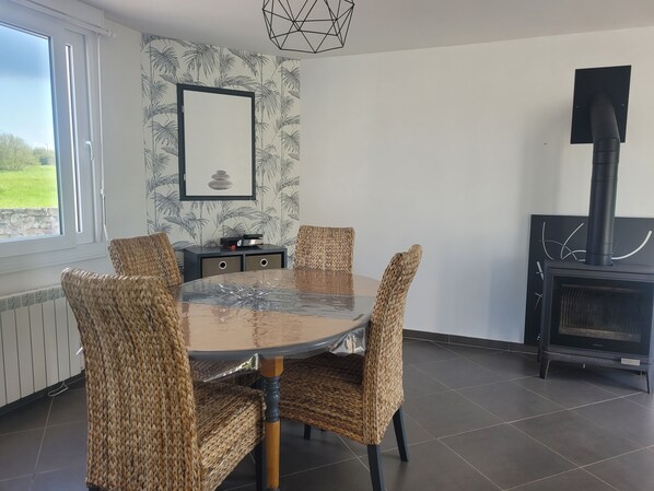 Spiseområde med et rundt glasbord, fletstole, et sort skab og en pejs.