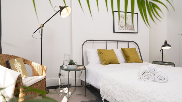 Un dormitorio ordenado con cama, mesitas de noche, una silla y una planta en maceta.