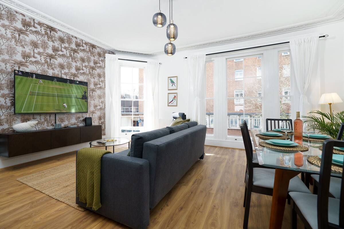 A modern living room with a large flat-screen TV, a sectional sofa, a dining table set for four, and a view of the outside through large windows.