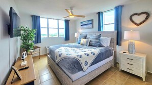 Een slaapkamer met een bed, nachtkastjes, een televisie, een plafondventilator en een raam met gordijnen.