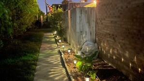 Un estrecho sendero de hormigón bordeado por una valla de madera y un muro de ladrillo, con pequeñas luces a lo largo del camino.