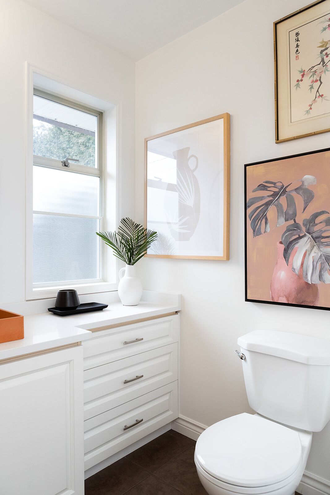 Ein modernes Badezimmer mit einem weißen Waschtisch, einem Fenster, einer Toilette und zwei gerahmten Kunstwerken.
