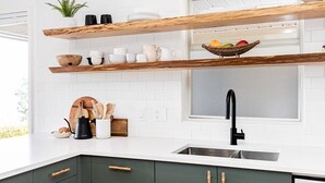 Una cocina con encimeras blancas, armarios verde oscuro, estantes de madera y un grifo negro.