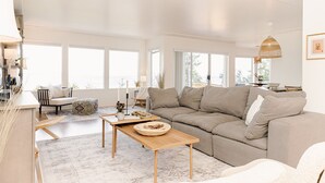 Un salón amplio con un sofá beige grande, una mesa de centro de madera y vistas al exterior a través de amplias ventanas.