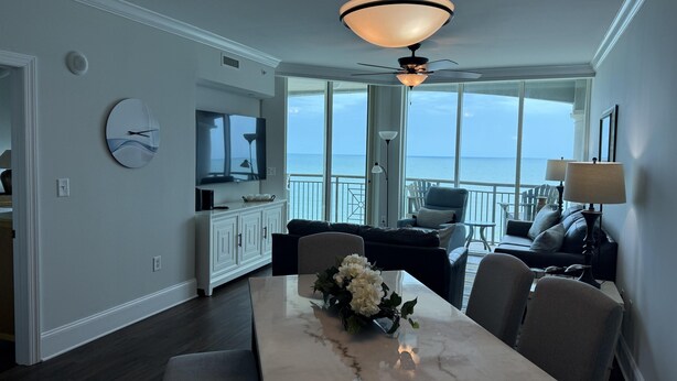 A modern living room with a large TV, a dining table with chairs, a sofa, and a view of the ocean. - MVG 1512 Oceanfront, Penthouse (North Myrtle Beach)