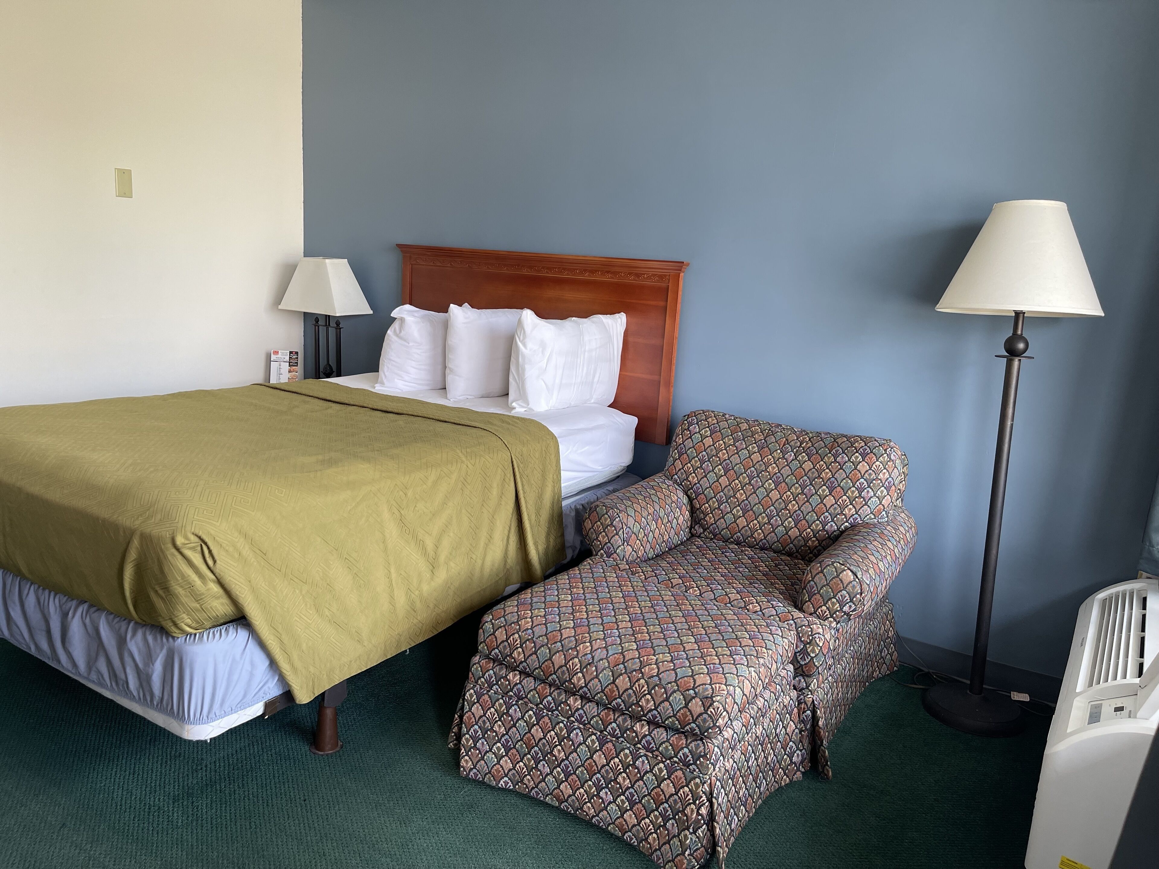 Een hotelkamer met een bed, een gestreept fauteuil, een staande lamp en een verwarming.