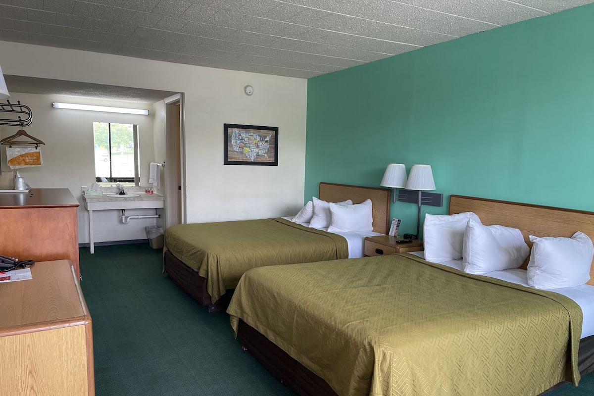 A hotel room with two beds, a desk, a mirror, a sink, and a framed picture on the wall.