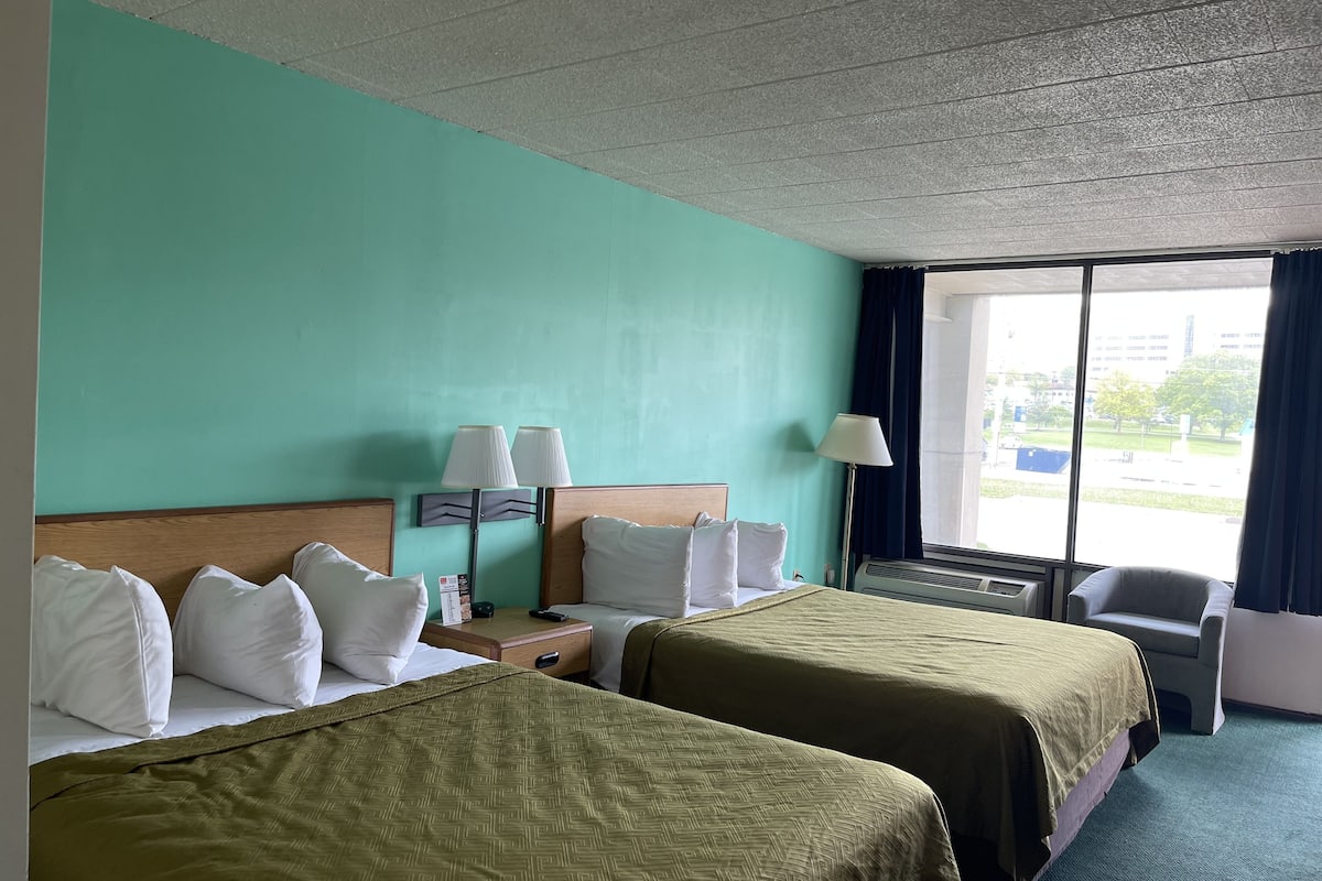 A hotel room with two beds, a desk, a chair, a window with curtains, and a view of the outside.