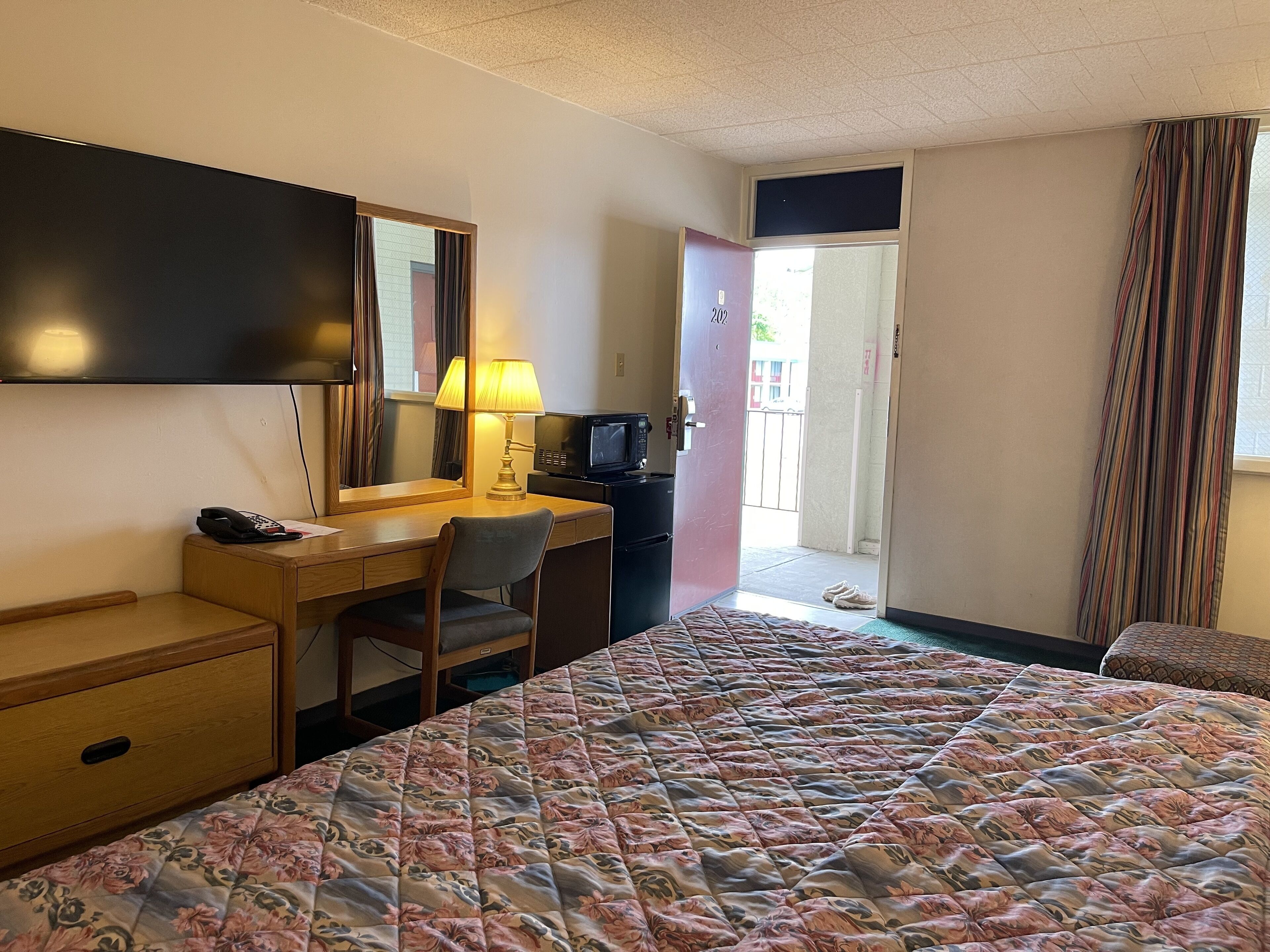 Une chambre d’hôtel comprenant un lit, un bureau avec une chaise, une télévision, une lampe et une porte donnant sur un balcon.