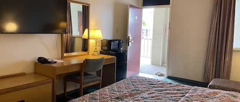 A hotel room with a bed, a desk with a chair, a TV, a lamp, and a door leading to a balcony.