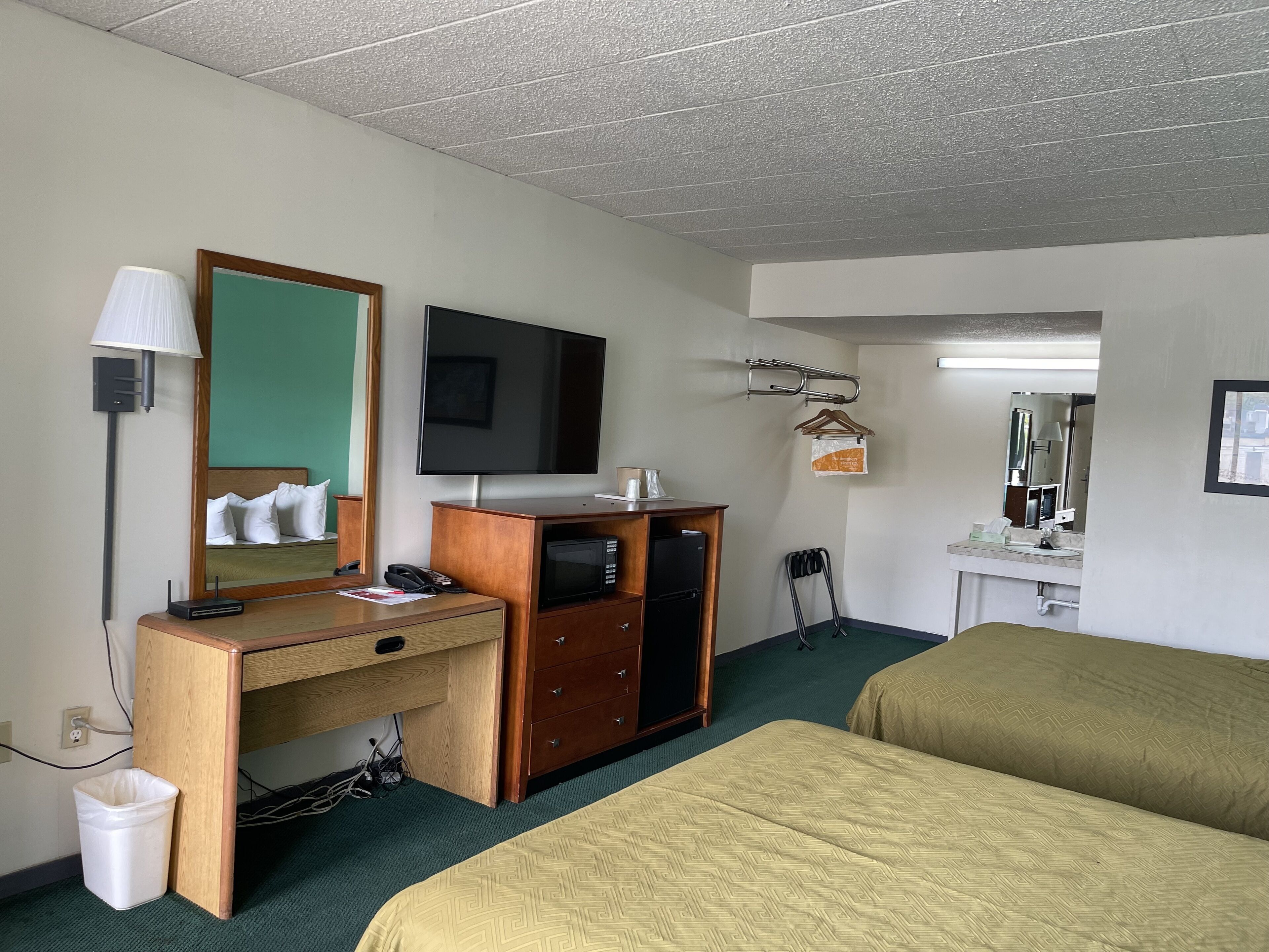 Habitación de hotel con escritorio de madera, televisor de pantalla plana, cama con ropa de cama verde, mesita de noche pequeña, cesto de basura y espejo.