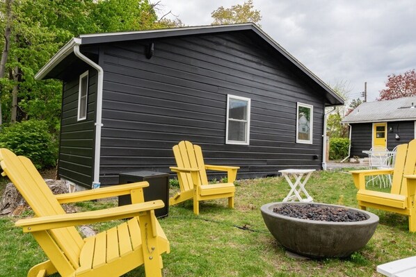 A black wooden cabin with a small deck, yellow Adirondack chairs, a fire pit, and a white table. - Double the Fun: Two Houses, Shared Yard & Fire Pit (Kingston)