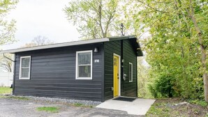 A small, single-story building with a dark exterior and a yellow door, surrounded by trees and a gravel driveway. - Double the Fun: Two Houses, Shared Yard & Fire Pit (Kingston)