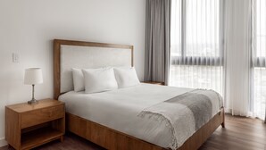 A neatly made bed with a wooden headboard and white bedding, a bedside table with a lamp, and a view of the outdoors through sheer curtains. - Master Suite DLX (Heredia)