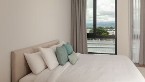 A bedroom with a large bed, a nightstand, and a window with a city view. - Master Suite DLX (Heredia)