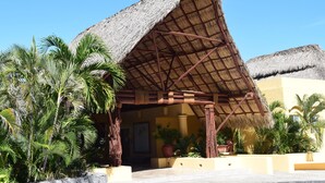 Un bâtiment d’hôtel de villégiature avec un toit de chaume, entouré de palmiers et de plantes en pot.