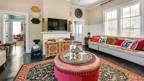 A spacious living room with a large sofa, a round ottoman, a flat-screen TV, and a decorative rug. - Grand Historic Home w/ Yard & Deck in High Point! (High Point)
