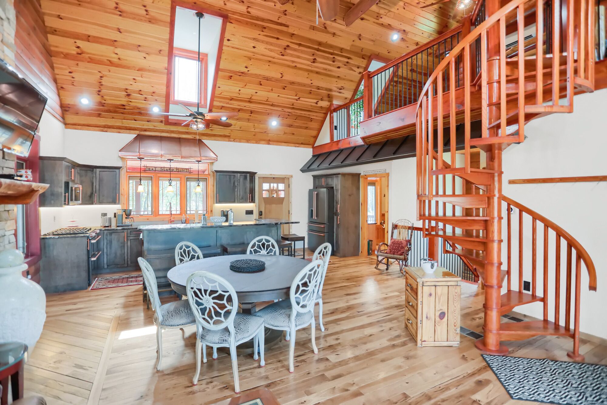Una cocina amplia con una isla central, un comedor con sillas blancas y una escalera de madera que conduce a un nivel superior.
