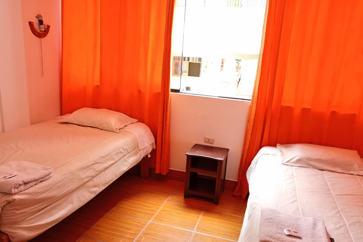 Une chambre avec deux lits, des rideaux orange, une petite table de chevet en bois et une fenêtre donnant sur des bâtiments.