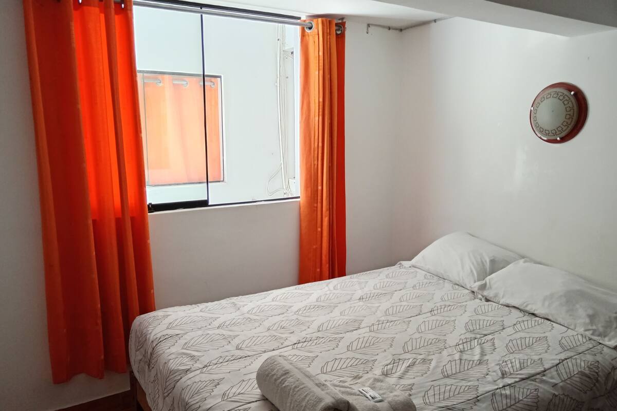 Une chambre à coucher avec un lit, des rideaux orange, une fenêtre et un miroir fixé au mur.