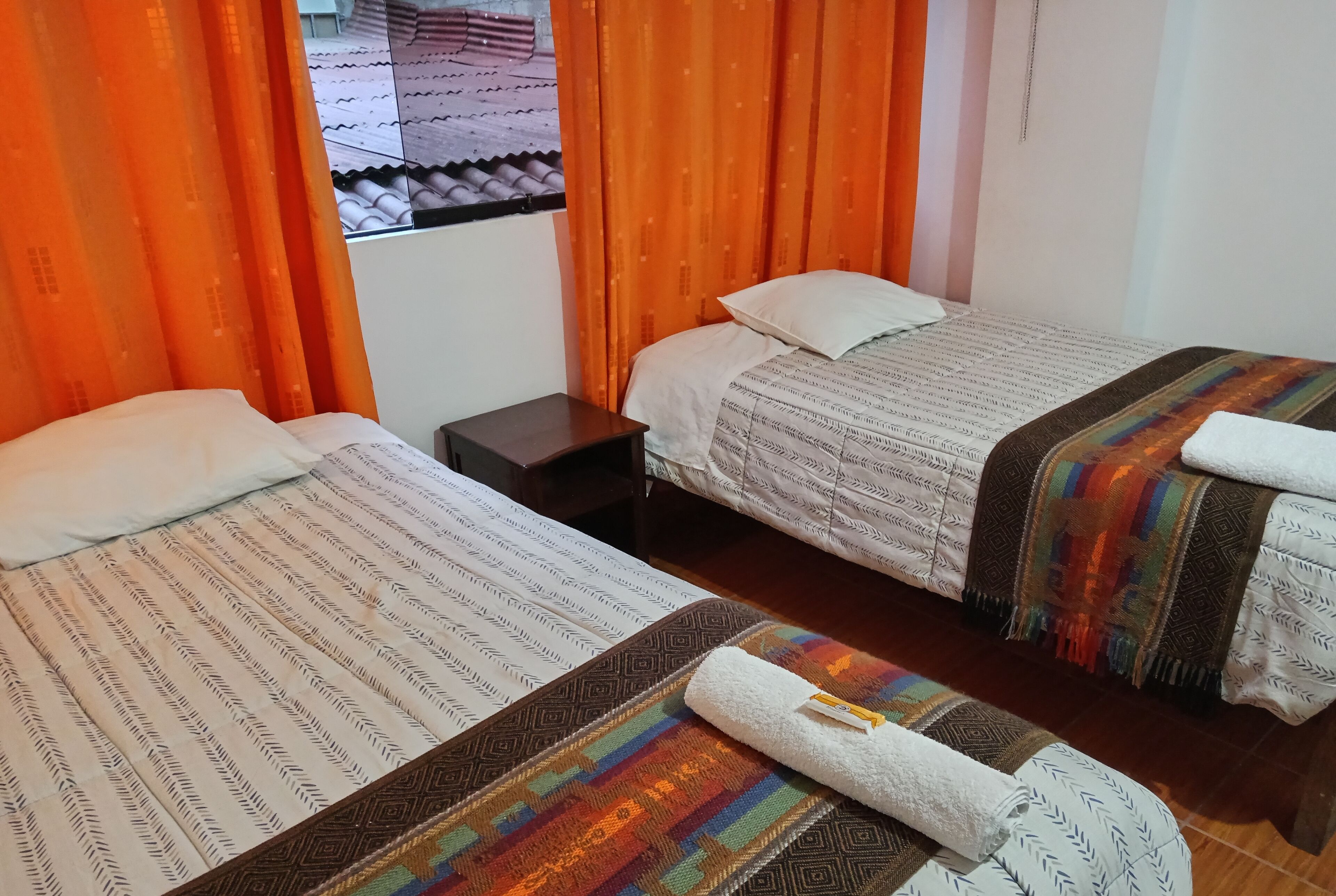 A hotel room with two beds, orange curtains, a small nightstand, and a window with a view of the outside.