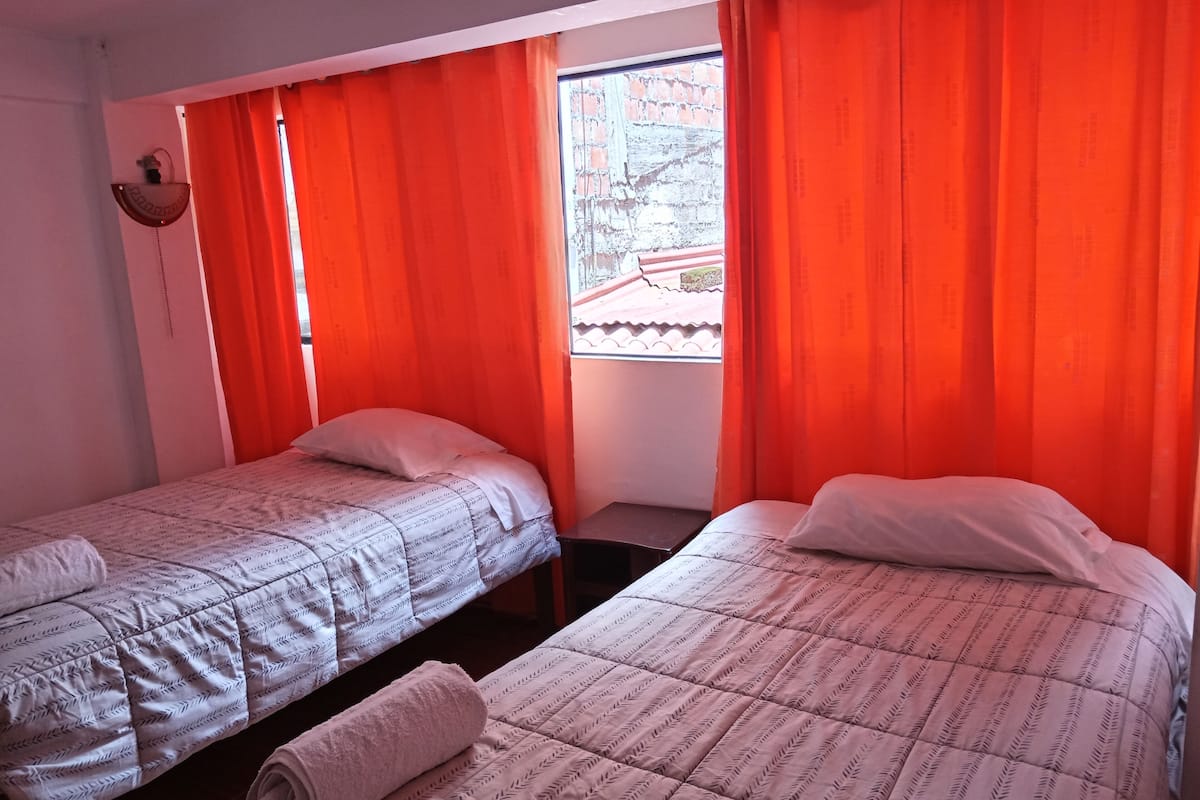 Une chambre avec deux lits simples, une petite table et une fenêtre avec des rideaux rouges.