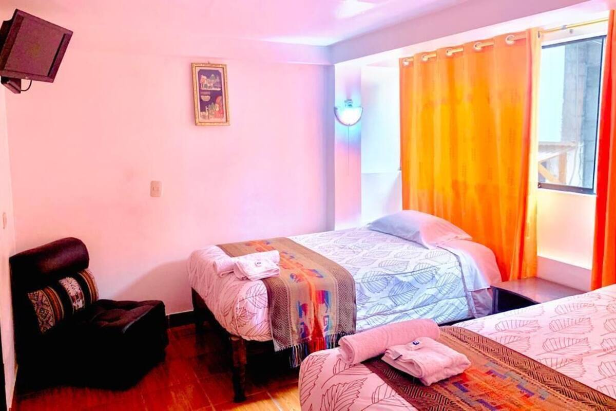 Une chambre d’hôtel avec deux lits, un téléviseur fixé au mur et une fenêtre avec des rideaux orange.