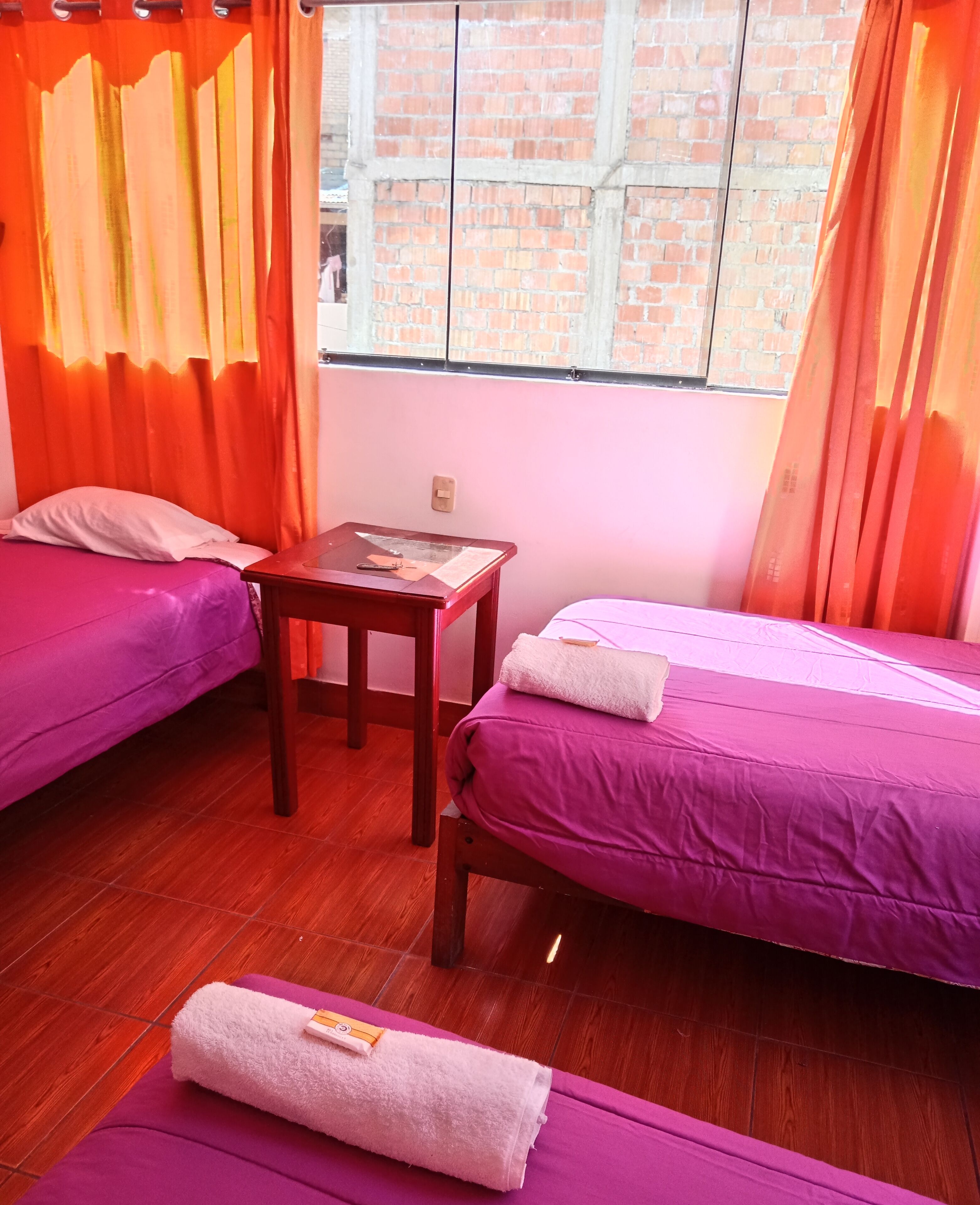 Una habitación con dos camas, una mesita y una ventana que da a paredes de ladrillo.