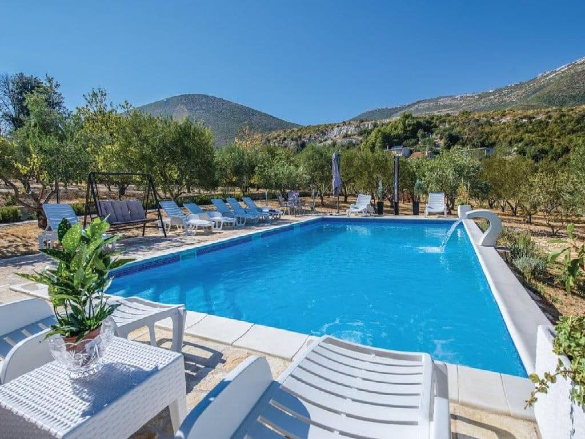 A rectangular swimming pool with white lounge chairs and umbrellas, surrounded by greenery and hills in the background.
