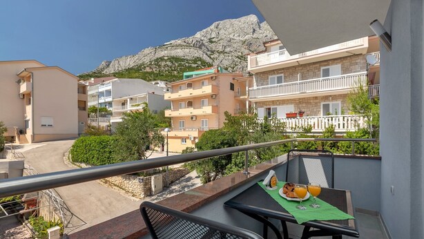Un balcón con una mesa lista para el desayuno, con vistas a una calle y edificios, y una montaña al fondo.