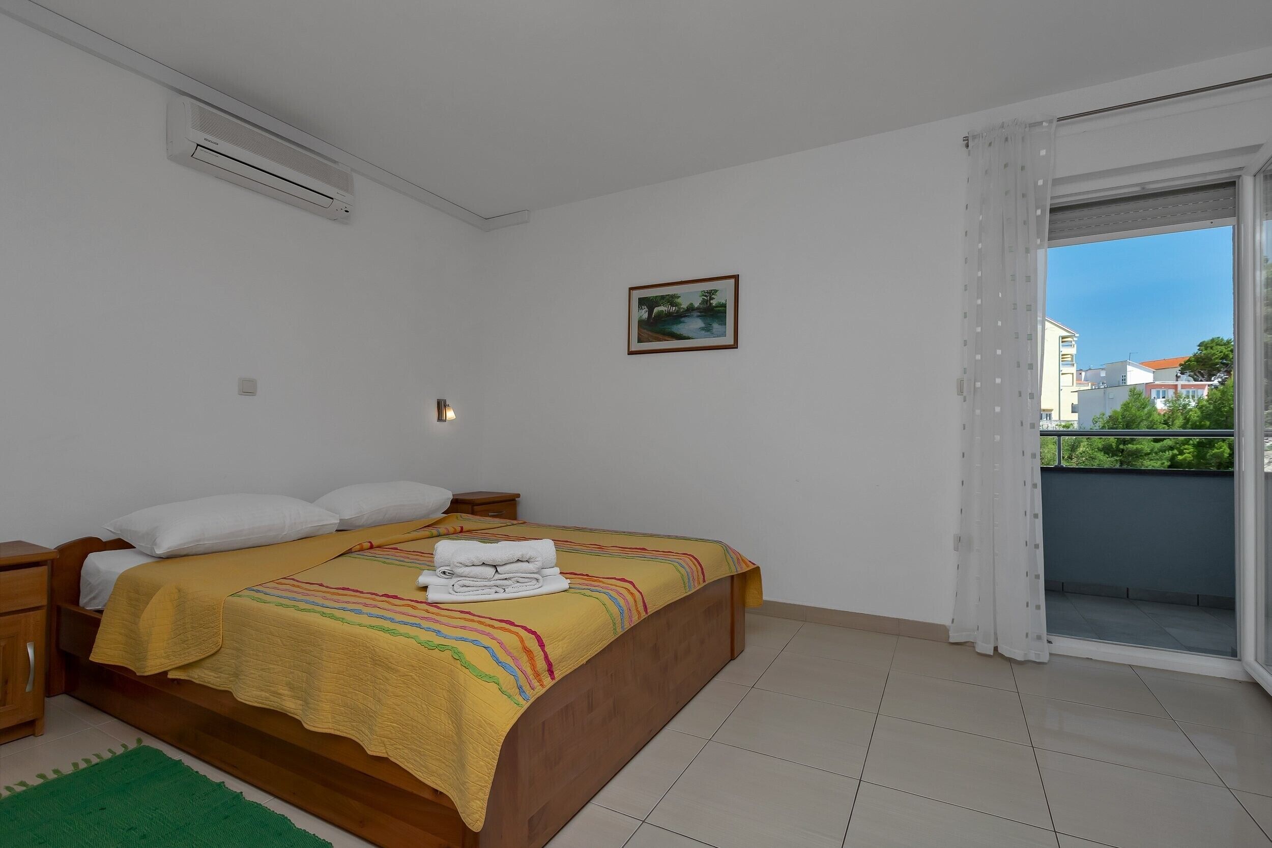 Un dormitorio con cama, mesita de noche, alfombra verde, un cuadro en la pared y vistas a edificios a través de una ventana.