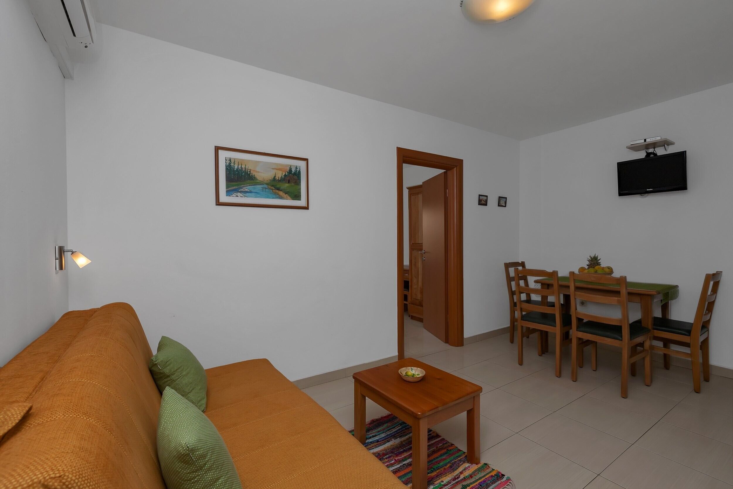 A living room with a sofa, a small table with a bowl, a dining table with chairs, a painting on the wall, and a TV mounted on the wall.