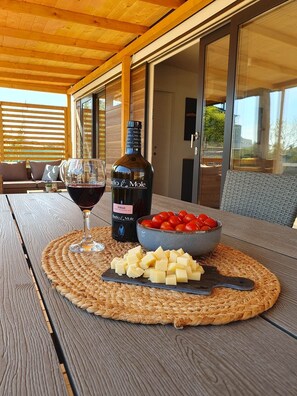 Une table en bois, sur laquelle on a posé un set de table en fibres tressées, une bouteille de vin Syrah, un verre de vin, un bol de tomates cerises et une planche à découper avec des cubes de fromage.