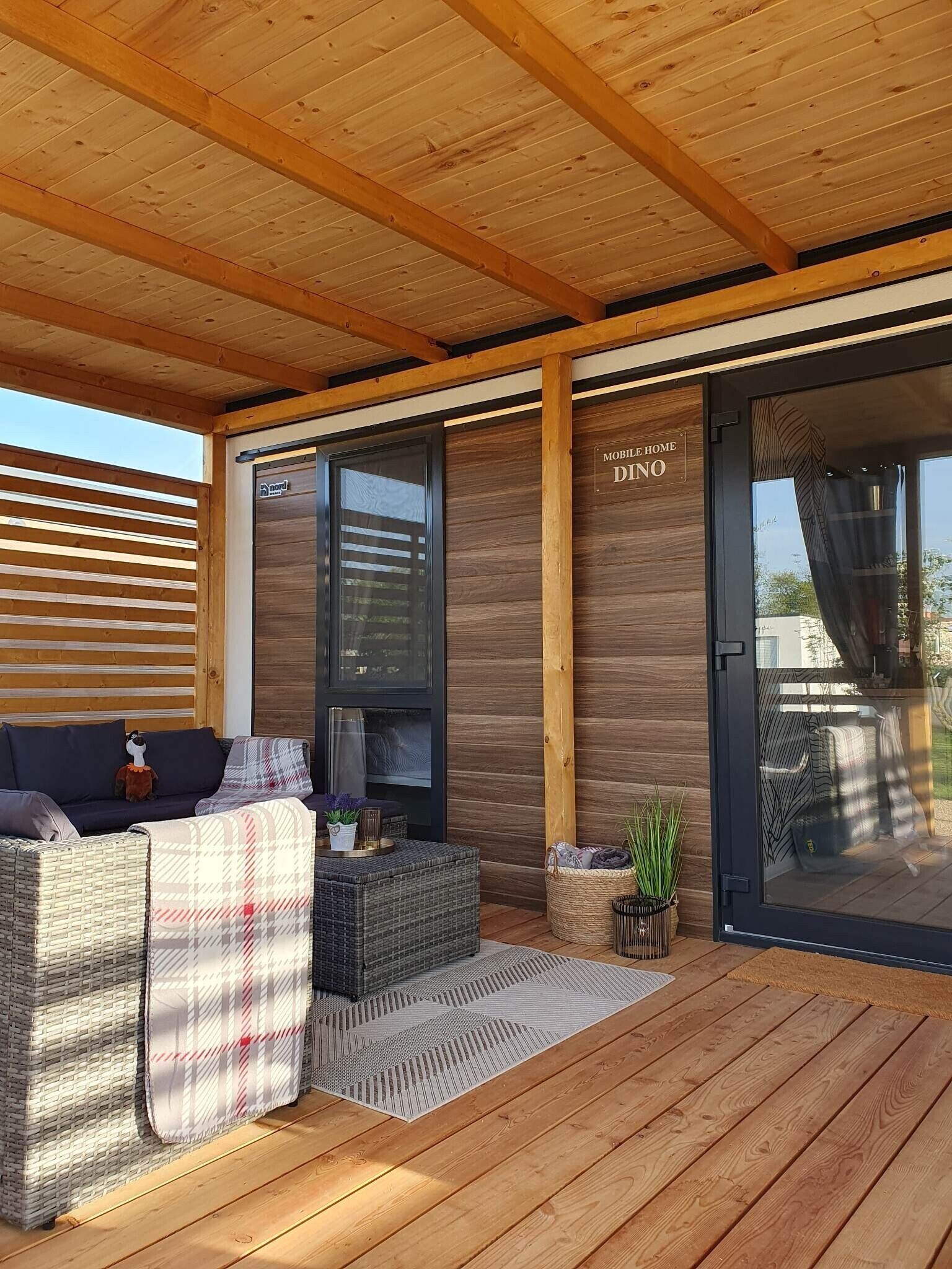A covered outdoor seating area with wicker furniture, a small table with a plant, and a sliding glass door labeled "MOBILE HOME DINO".