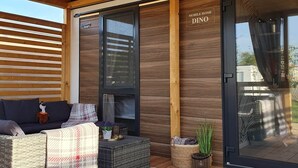 Un espace extérieur aménagé, abrité et doté de mobilier en osier, d’une petite table avec une plante et d’une porte-fenêtre coulissante portant l’inscription « MOBILE HOME DINO ».