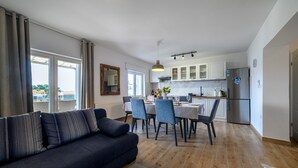 Un soggiorno moderno con un divano scuro, zona pranzo con sedie e tavolo, cucina con pensili bianchi e frigorifero in acciaio inossidabile.
