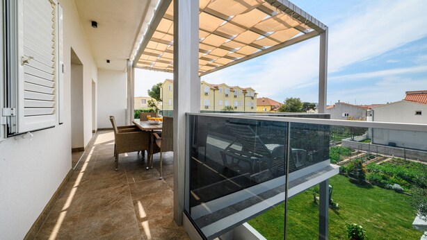 Un balcone con una porta a vetri, un tavolo da pranzo con sedie e vista su un giardino e edifici sullo sfondo.