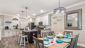 A modern kitchen with white cabinets, a central island, and bar stools. There is a dining table set with colorful plates and glasses. - Screened pool+Climate controlled game room+Outdoor TV+Fire pit+ Near the beach! (Navarre)