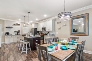 A modern kitchen with white cabinets, a central island, and bar stools. There is a dining table set with colorful plates and glasses. - Screened pool+Climate controlled game room+Outdoor TV+Fire pit+ Near the beach! (Navarre)